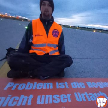 Mensch in oranger Weste sitzt auf Rollbahn, vor ihm ein Plakat mit der Aufschrift 