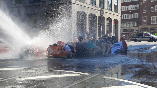 Eine Gruppe von Unterstützer*innen der Letzten Generation sitzt auf einer nassen Straße, während ein Wasserwerfer der Polizei im Hintergrund zu sehen ist. 