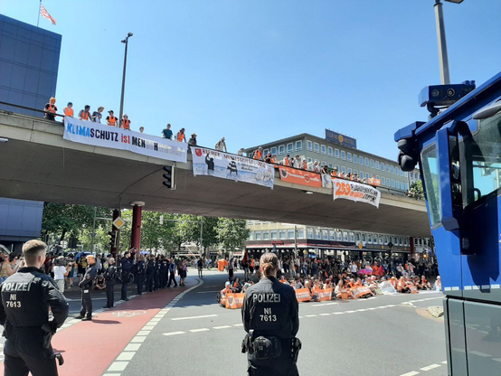 Polizisten und Unterstützer*innen der Letzten Generation versammeln sich an einer Kreuzung unter einer Brücke. Auf der Brücke stehen weitere Protestierende mit Bannern, die auf Klimaschutz hinweisen.