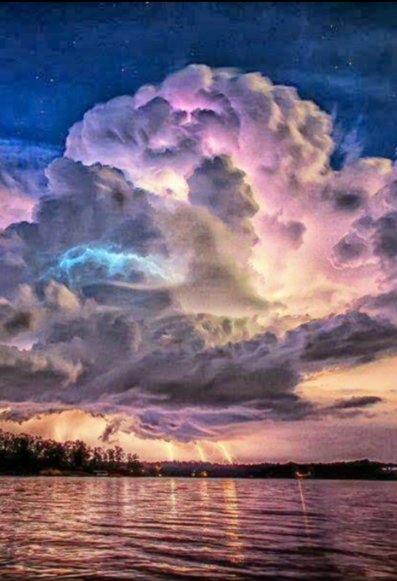 Storm clouds over a bay with tree covered land in the background. Flashes of lightening emanating from the bottom of the clouds light up the ground and water, with some lightening farther up towards the top of the clouds. The clouds are colored in shades of white, shades of purple and shades of grey.