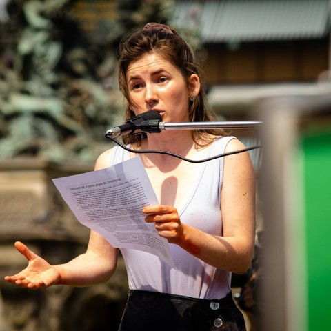 Person steht mit einem Zettel vor einem Mikrofon und hält eine Rede