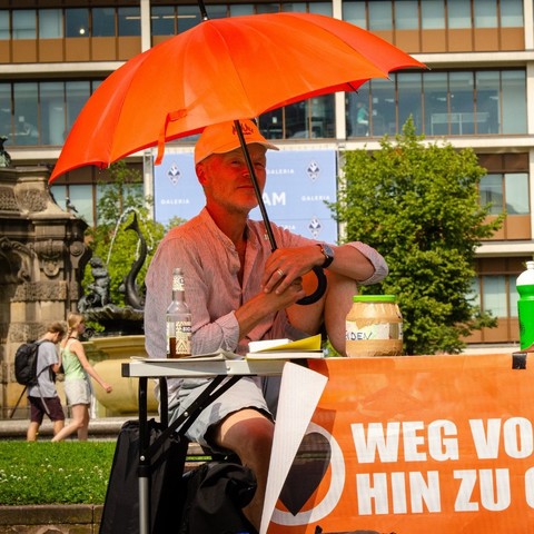 Person sitzt unter einem orangenen Regenschirm vor einem Banner der Letzten Generation