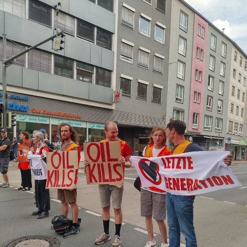 Einige Menschen stehen in auf einer Straße und halten Schilder und Banner hoch. Auf ihnen steht Letzte Generation und OIL KILLS