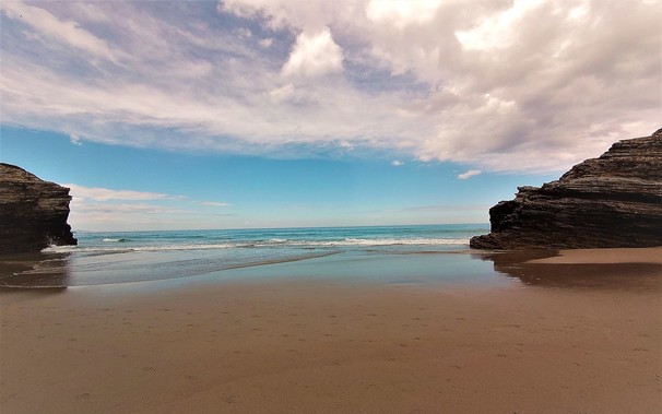 In the foreground, a stretch of flat sandy beach, still wet from the waves, with footprints visible. The beach is framed by shale cliffs on the left and right, and beyond that is the blue-green ocean with a bright blue sky and some white clouds above.