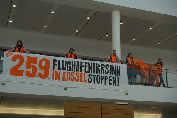 Das Banner hängt im Flughafen und Menschen in orangenen Warnwesten stehen dahinter