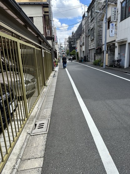 A narrow street in a city with a sidewalk on the left, lined with a golden fence. Parked cars are visible in a garage, but not on the street, and a person walks in the distance. The sky is partly cloudy.