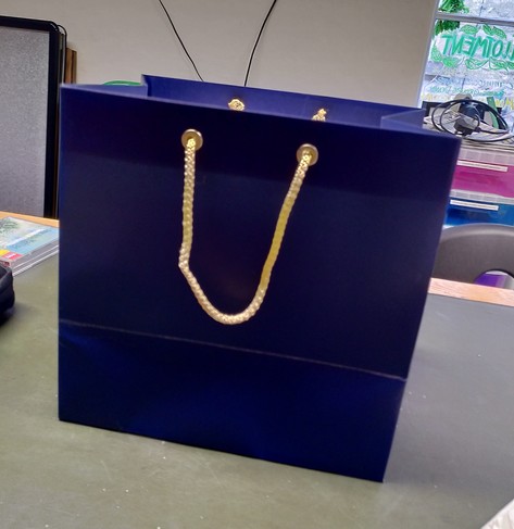 Image of a large dark blue paper bag with gold braid handles on a large green table. 